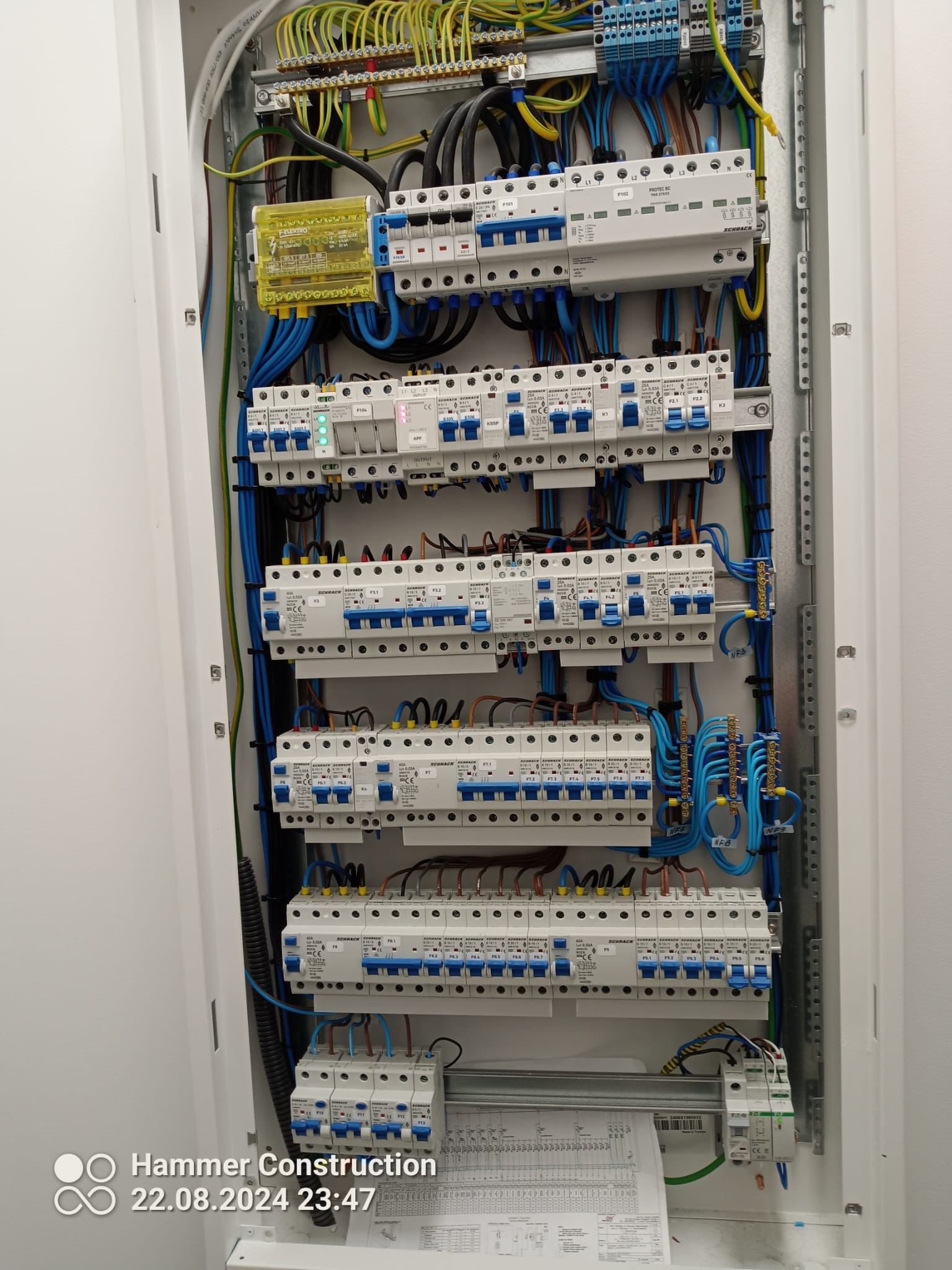 Rozdzielnica elektryczna z widocznymi zabezpieczeniami, przewodami w kolorach niebieskim, żółto-zielonym i brązowym, zamontowana w białej obudowie, z dokumentacją techniczną na dole.