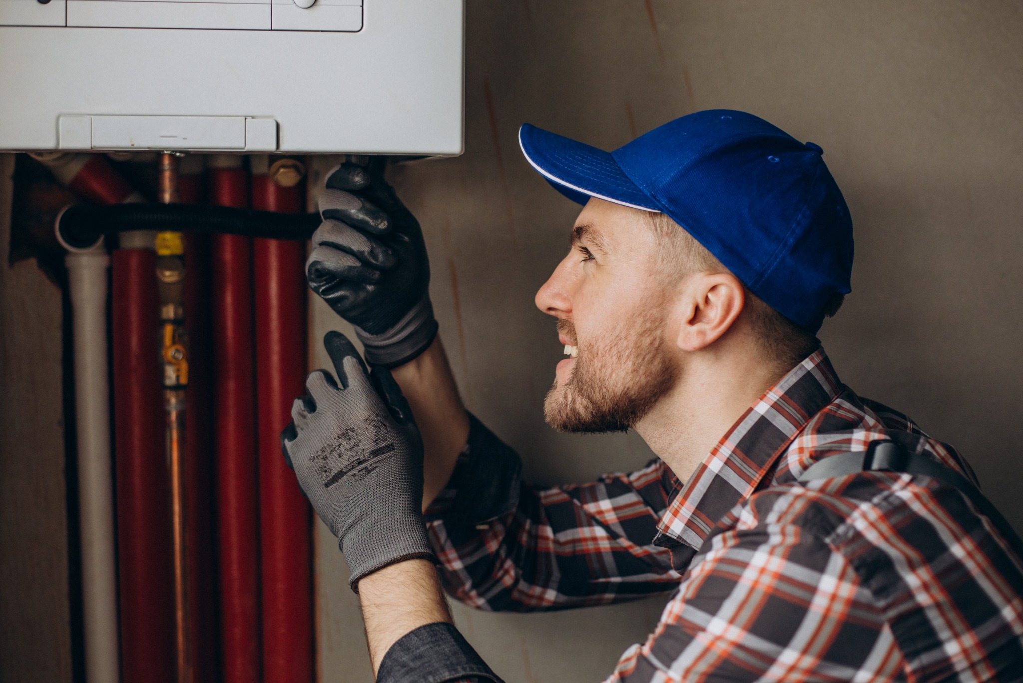 Instalator w niebieskiej czapce i roboczych rękawicach przyłącza rurę do białego pieca gazowego, widoczne czerwone rury instalacji.