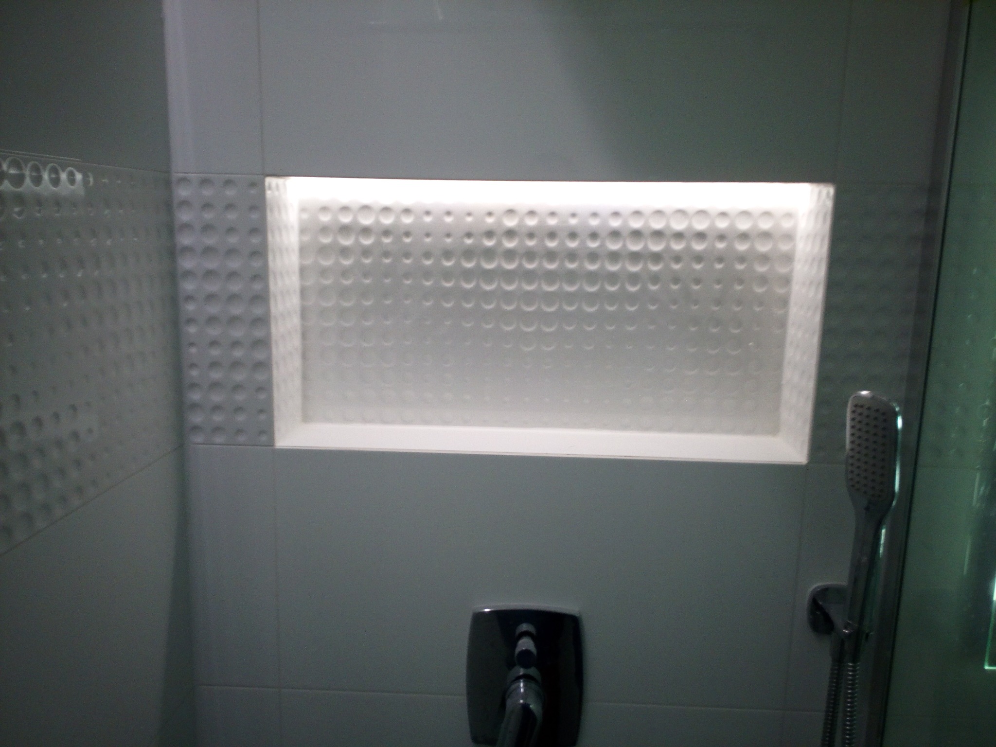 Wnęka w ścianie łazienki wyłożona białą, wzorzystą glazurą, podświetlona od wewnątrz, z widoczną baterią prysznicową i słuchawką.