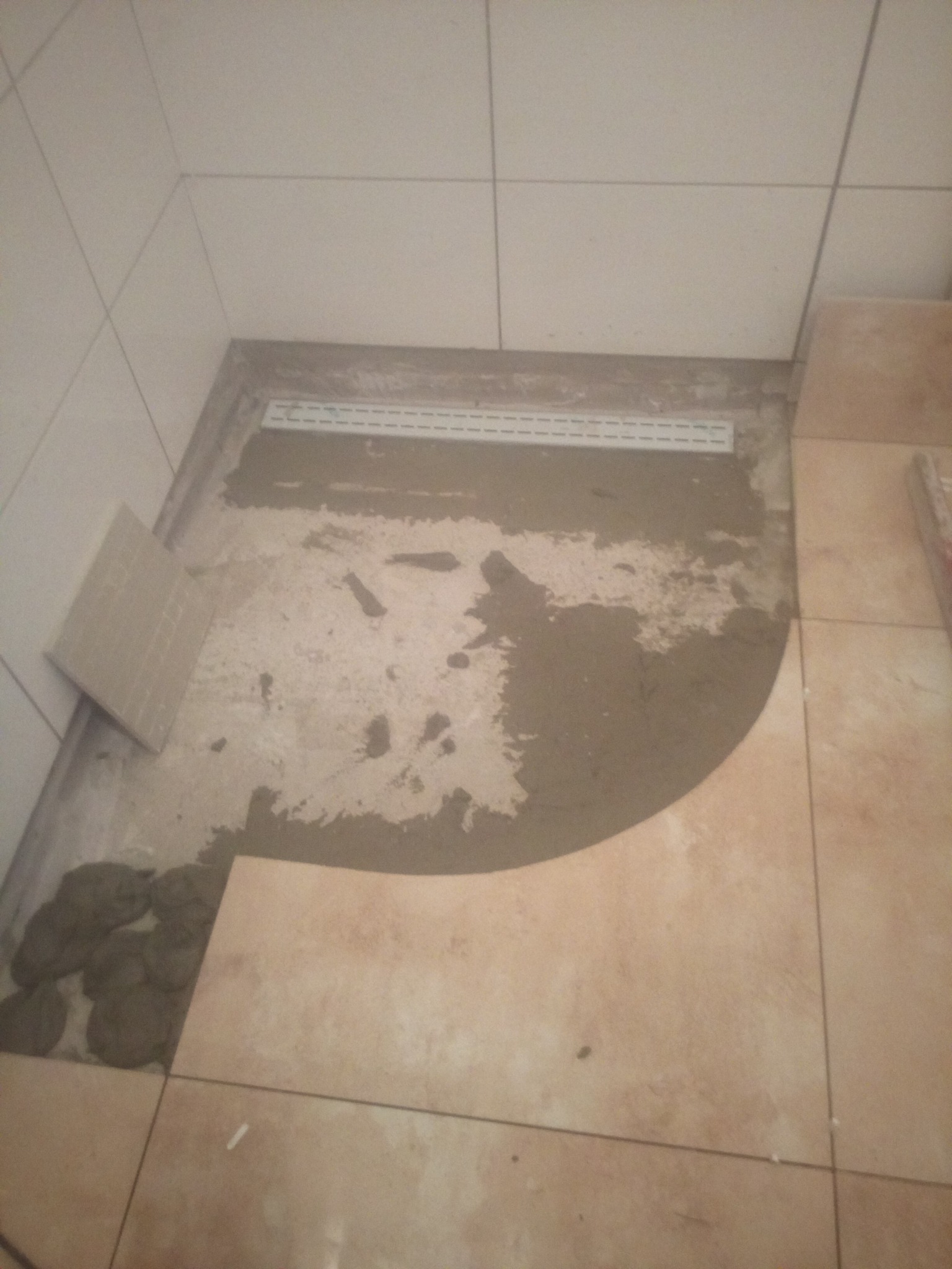 Niedokończona podłoga w łazience z ułożonymi płytkami w rogu i widocznym odpływem liniowym w wylewce.