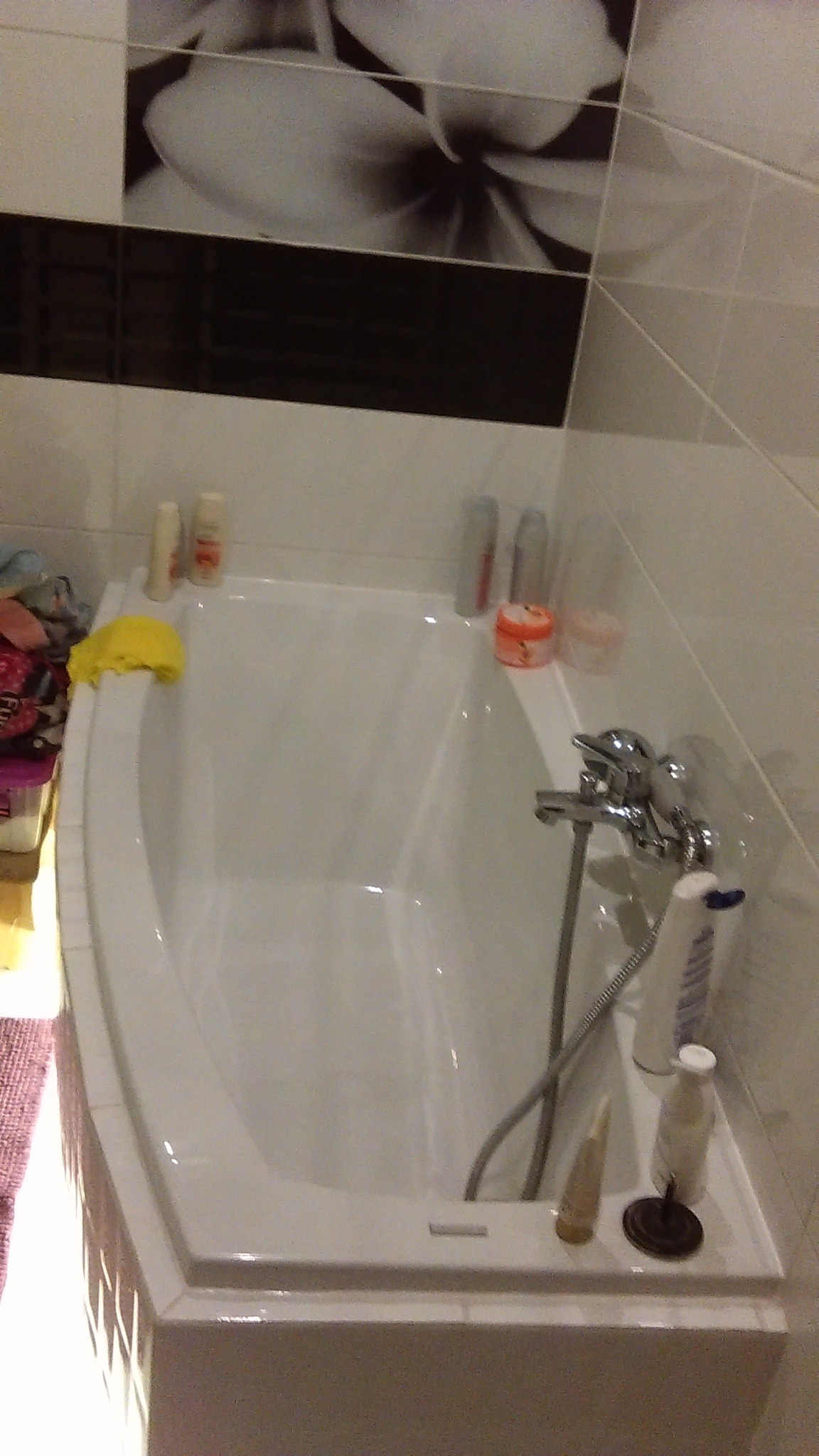 Biała wanna z chromowaną baterią i prysznicem w wykafelkowanej łazience, nad wanną widoczne kosmetyki i dekoracyjny motyw kwiatowy na płytkach