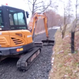 Pomarańczowa minikoparka na gąsienicach, profil, na żwirowej ścieżce w terenie leśnym, zima, widoczne drzewa bez liści i resztki śniegu.