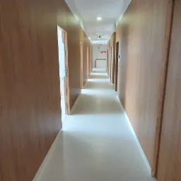 Długi korytarz z drewnianymi panelami ściennymi i drzwiami, jasna podłoga, widoczny znak ewakuacyjny w oddali.