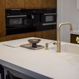 MEBROLL - TOMASZ OSZUST - Nowoczesna kuchnia z drewnianymi szafkami, białym blatem i złotą armaturą. Na blacie dekoracje: misa, kwiaty i radio. W tle wbudowane sprzęty AGD.