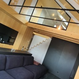 MEBROLL - TOMASZ OSZUST - Nowoczesne wnętrze z drewnianą zabudową, antresolą i czarną podłogą. Widoczny fragment sofy i stolika z dekoracyjnym wzorem. Nietypowy kąt ujęcia.