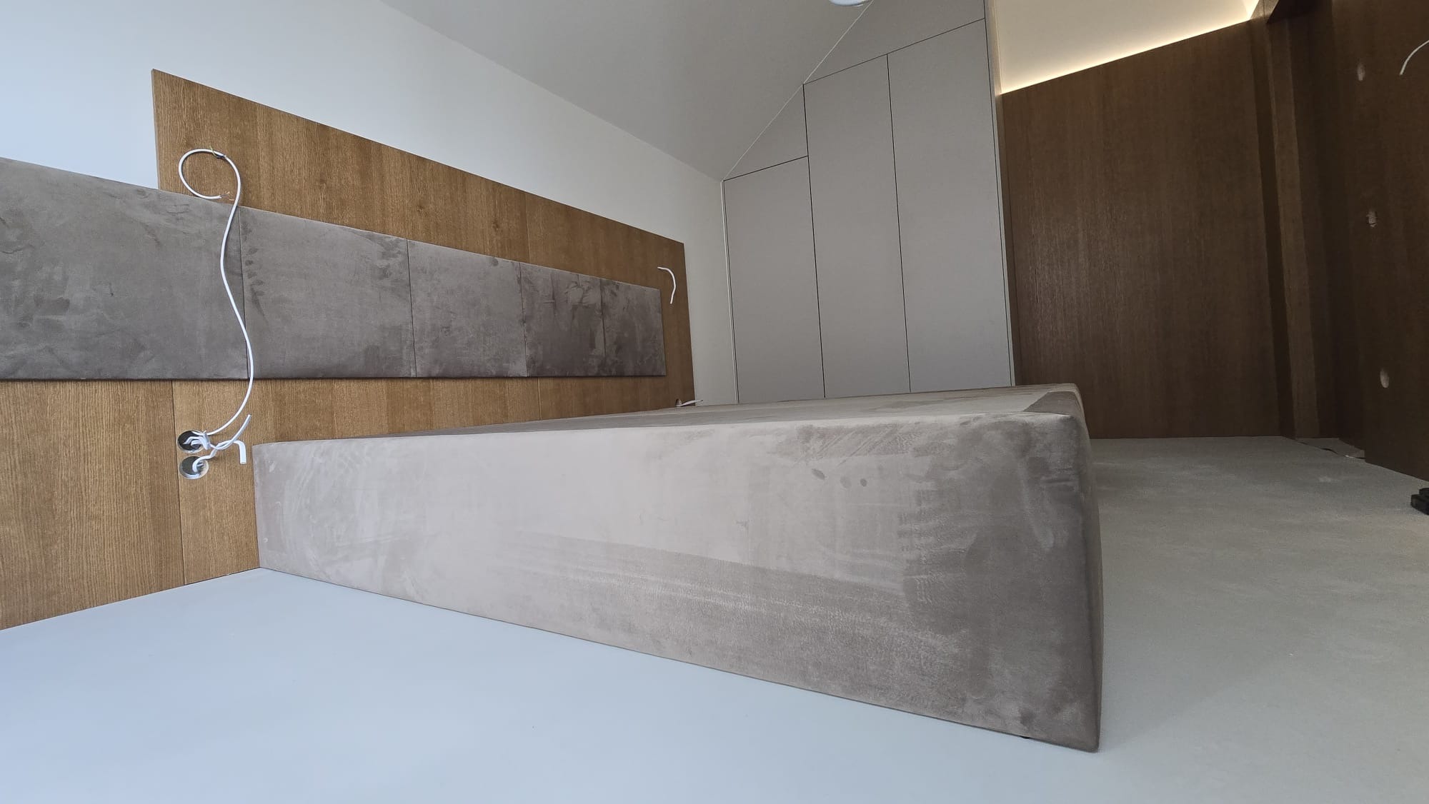 Wnętrze sypialni w trakcie wykańczania: wezgłowie z paneli tapicerowanych i drewnianych, zabudowa szafy wnękowej, widoczne kable elektryczne. Minimalistyczny design.