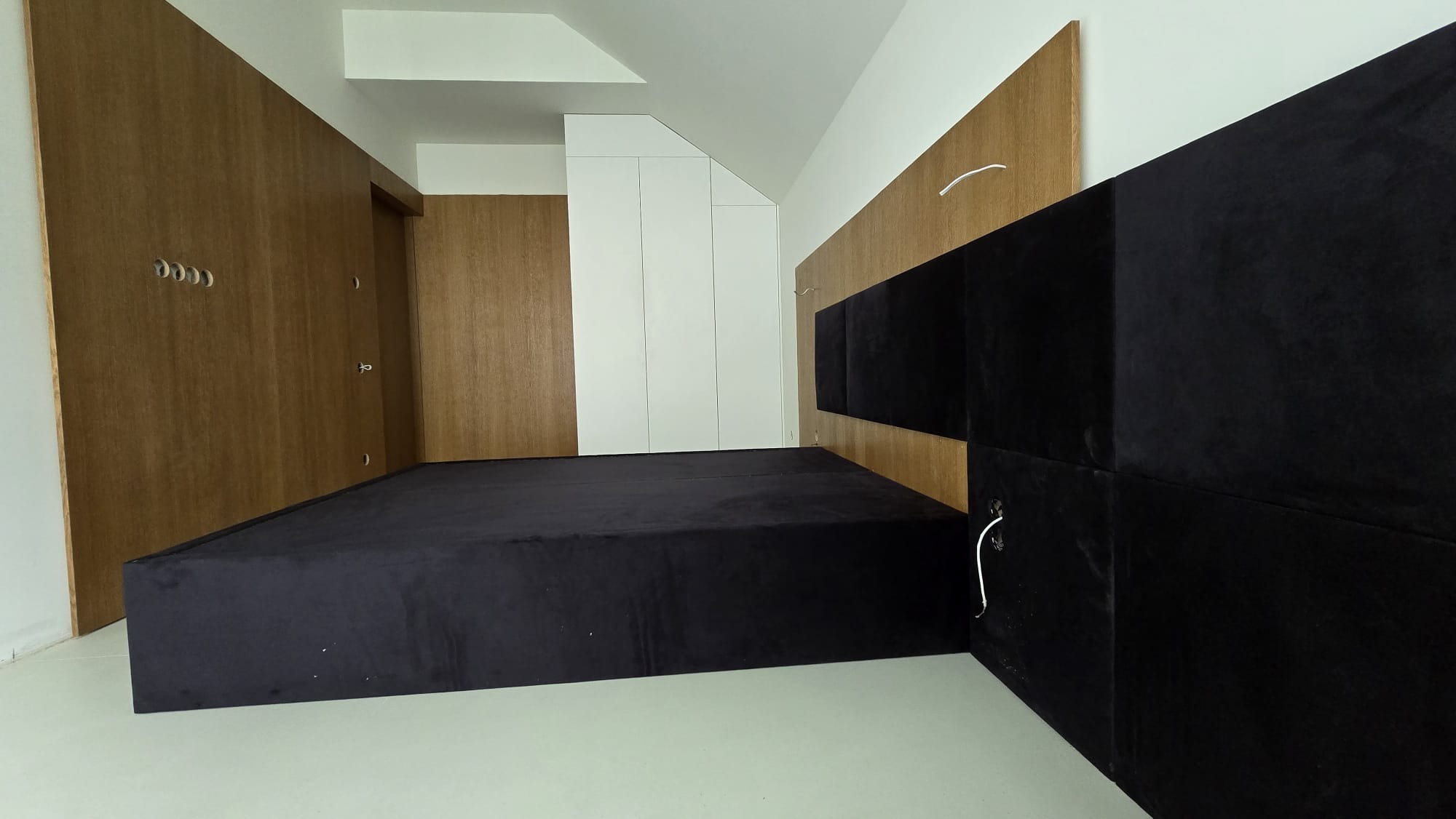 Nowoczesna sypialnia z czarnym, tapicerowanym łóżkiem i wezgłowiem z paneli. Drewniana zabudowa ściany z wieszakami i biała szafa w skosie.