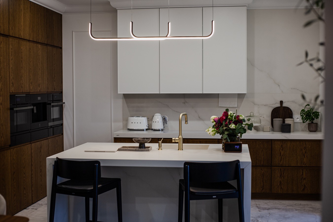 Nowoczesna kuchnia z wyspą, drewnianymi szafkami i białymi górnymi szafkami. Nad wyspą designerska lampa. Na blacie kwiaty, czajnik i toster.