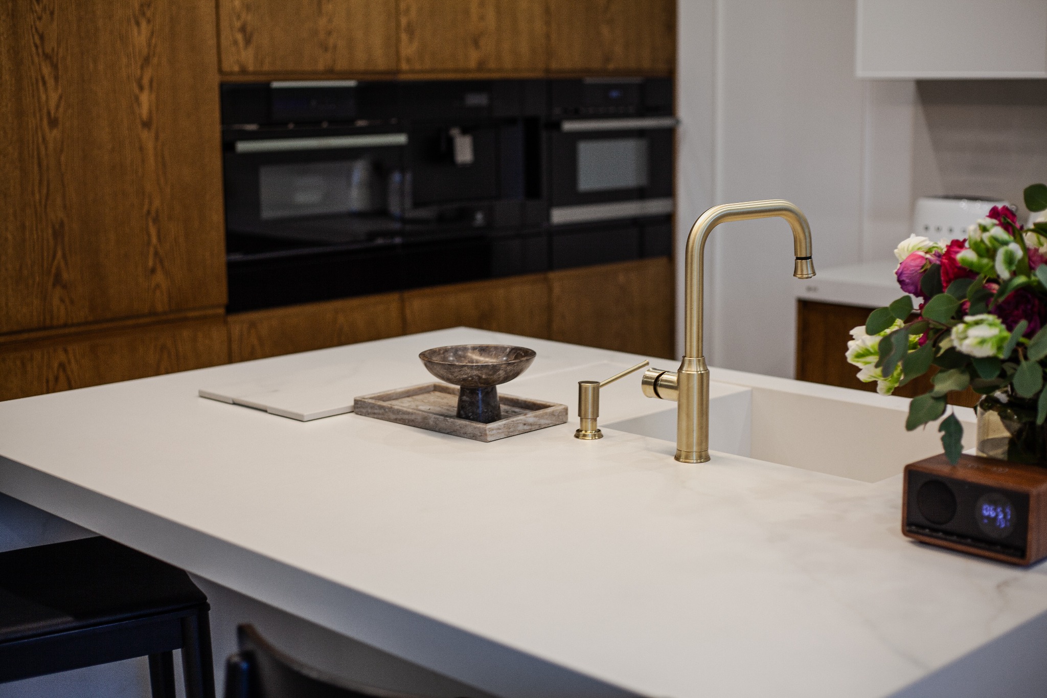 Nowoczesna kuchnia z drewnianymi szafkami, białym blatem i złotą armaturą. Na blacie dekoracje: misa, kwiaty i radio. W tle wbudowane sprzęty AGD.