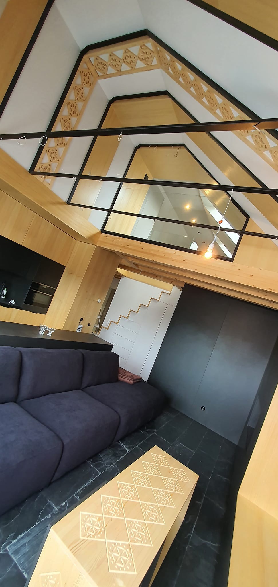 Nowoczesne wnętrze z drewnianą zabudową, antresolą i czarną podłogą. Widoczny fragment sofy i stolika z dekoracyjnym wzorem. Nietypowy kąt ujęcia.