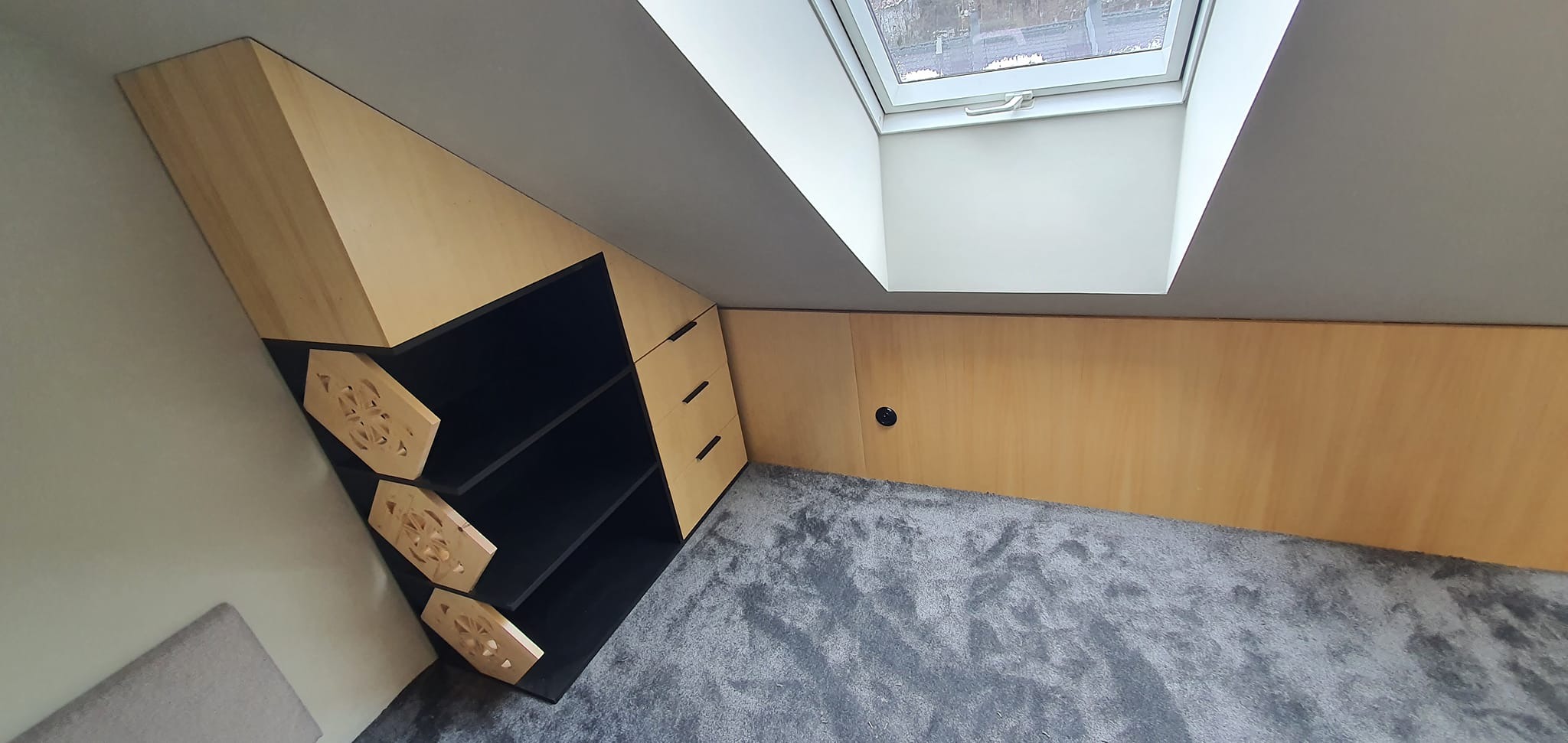 Funkcjonalna zabudowa pod skosem z jasnego drewna i czarnymi akcentami, z półkami i szafkami, dopasowana do przestrzeni poddasza z oknem dachowym.