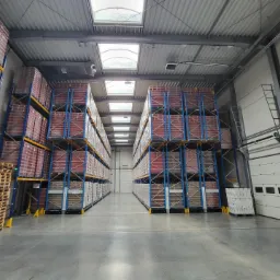 Pełny magazyn z wysokimi regałami, na których składowane są towary zapakowane w folię, oświetlony naturalnym światłem z okien dachowych.