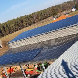 Hymon Fotowoltaika Oddział Gdańsk - Instalacja paneli słonecznych na dachu budynku gospodarczego z widocznym sprzętem rolniczym wewnątrz, w tle las i zabudowania.