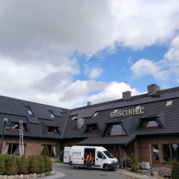 Hymon Fotowoltaika Oddział Gdańsk - Budynek Gościńca z panelami słonecznymi na dachu, drabina oparta o dach, biały bus firmy instalacyjnej z otwartymi drzwiami i pracownikiem w kamizelce odblaskowej.