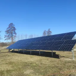 Instalacja paneli słonecznych na metalowej konstrukcji, widoczny na tle błękitnego nieba i drzew, z nietypowym, pomalowanym na różowo i szaro samochodem w tle po lewej stronie.