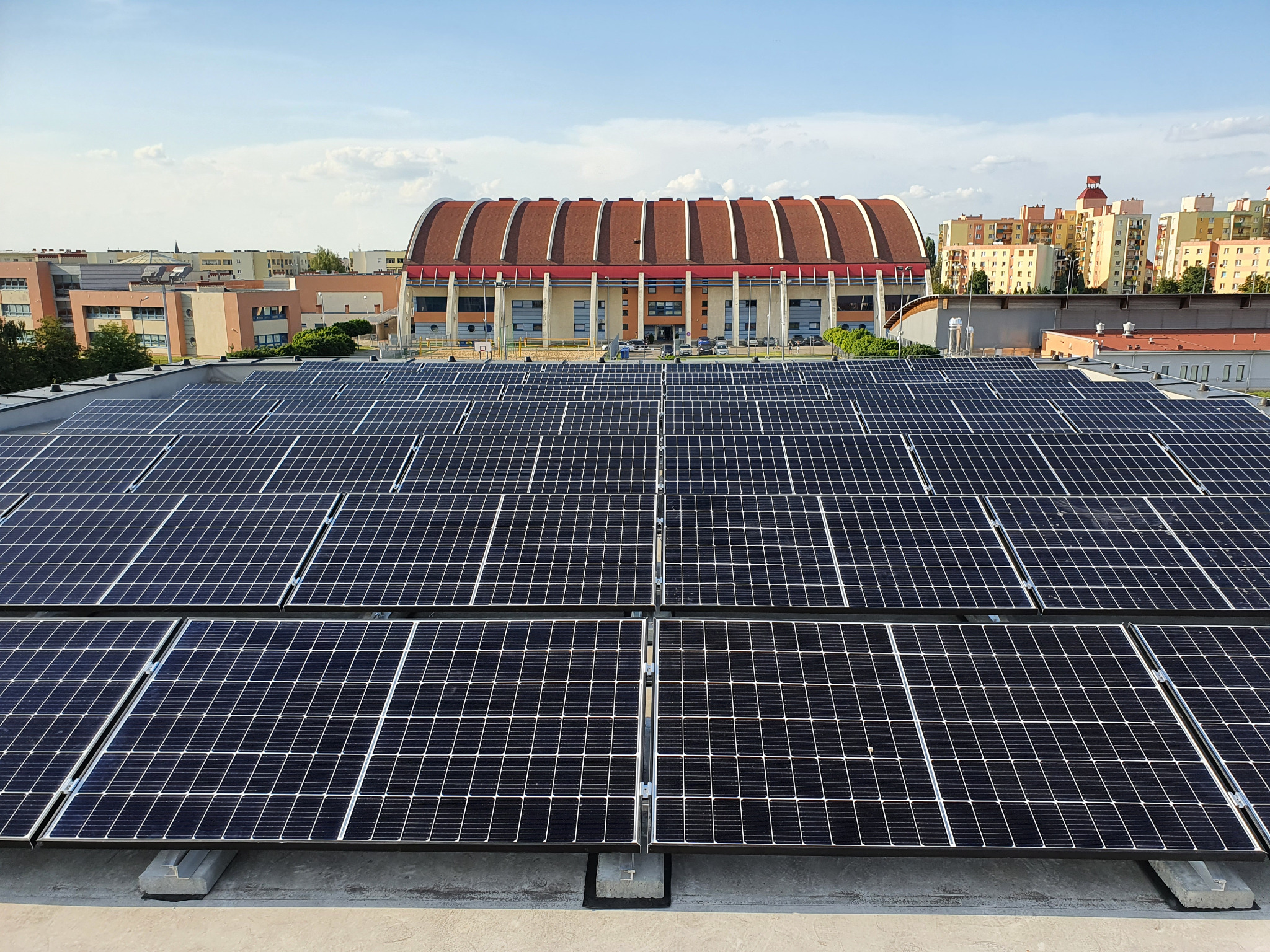 Rozległa instalacja fotowoltaiczna na dachu budynku z widokiem na halę sportową o charakterystycznym łukowym dachu i zabudowę miejską w tle.