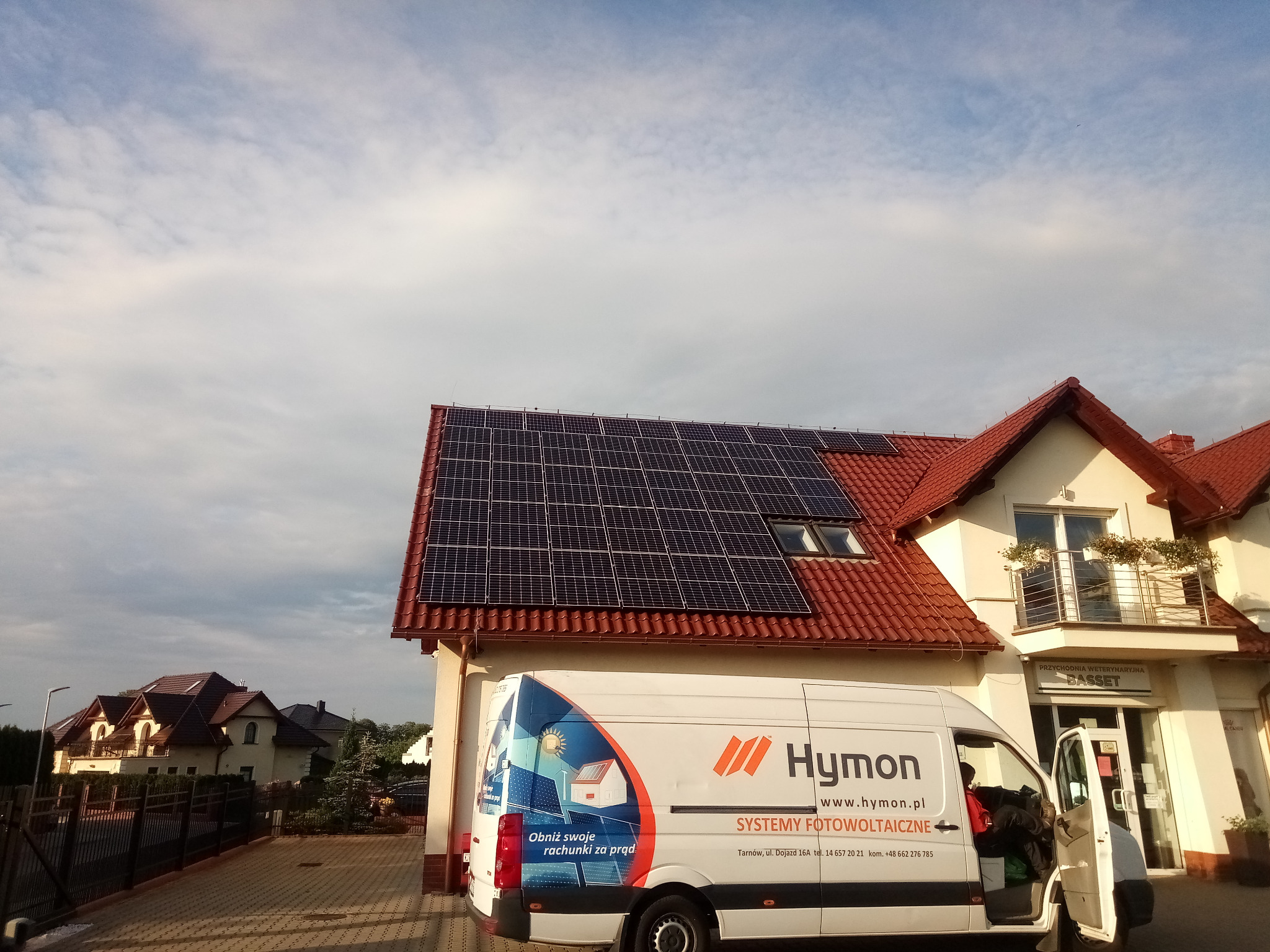 Dom z czerwoną dachówką i panelami słonecznymi na dachu, obok zaparkowany biały van z logo firmy instalującej systemy fotowoltaiczne, widoczny wjazd do przychodni weterynaryjnej.