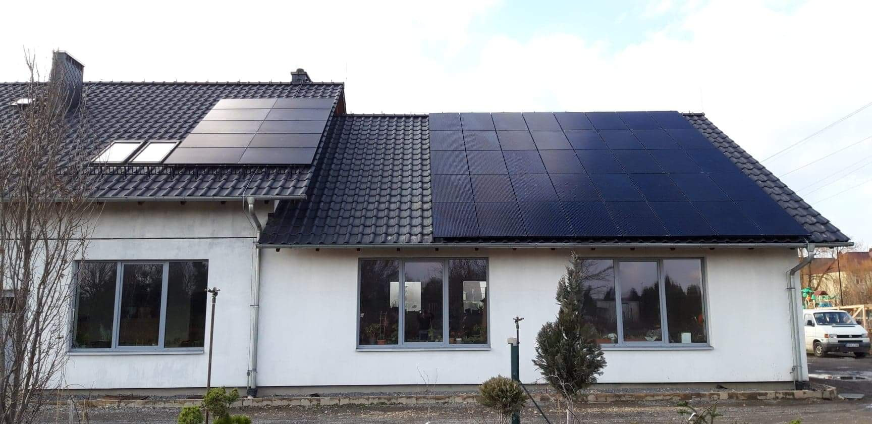 Czarny dach z panelami słonecznymi na białym domu jednorodzinnym, widoczne okna z szarymi ramami i fragment ogrodu.