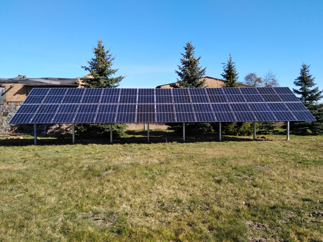 Rząd paneli słonecznych na metalowej konstrukcji, ustawiony na trawiastym terenie przed budynkiem z kamienną fasadą, widoczne wysokie drzewa iglaste, błękitne niebo.