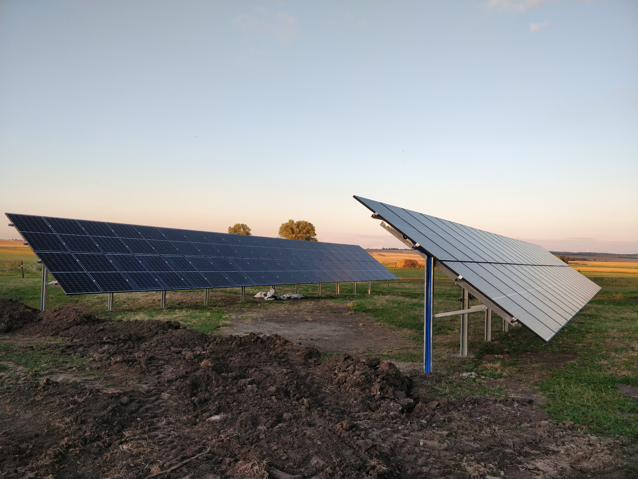 Dwa rzędy paneli słonecznych na polu, z widoczną świeżo przekopaną ziemią w pierwszym planie i zachodzącym słońcem w tle.