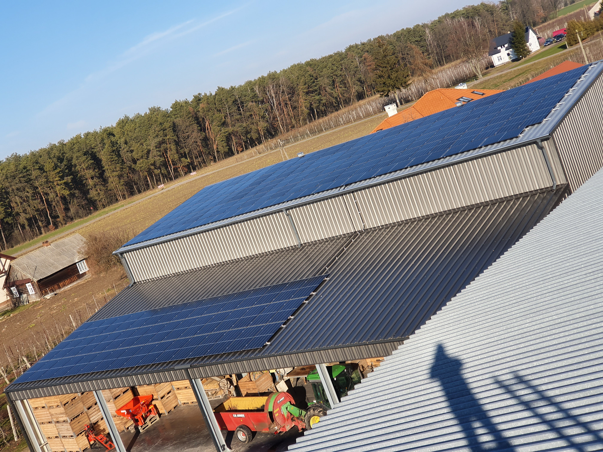 Instalacja paneli słonecznych na dachu budynku gospodarczego z widocznym sprzętem rolniczym wewnątrz, w tle las i zabudowania.