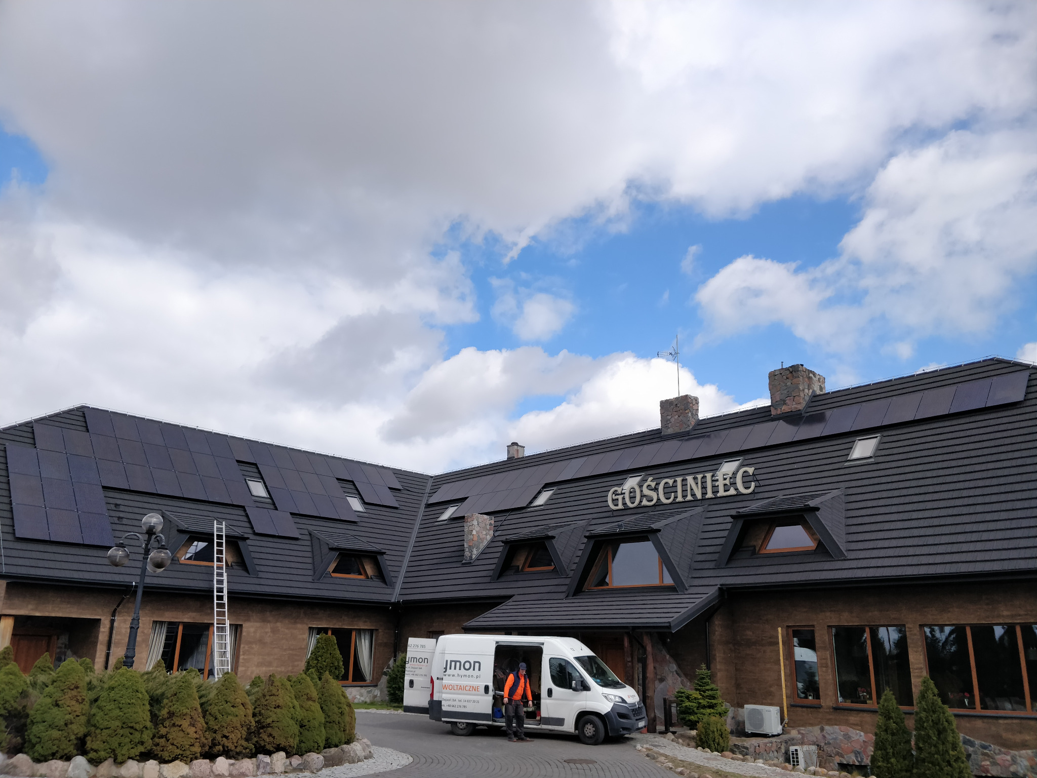 Budynek Gościńca z panelami słonecznymi na dachu, drabina oparta o dach, biały bus firmy instalacyjnej z otwartymi drzwiami i pracownikiem w kamizelce odblaskowej.