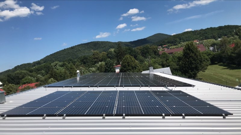 Panele fotowoltaiczne zamontowane na dachu z blachy trapezowej, w tle zalesione wzgórze pod błękitnym niebem z białymi obłokami.