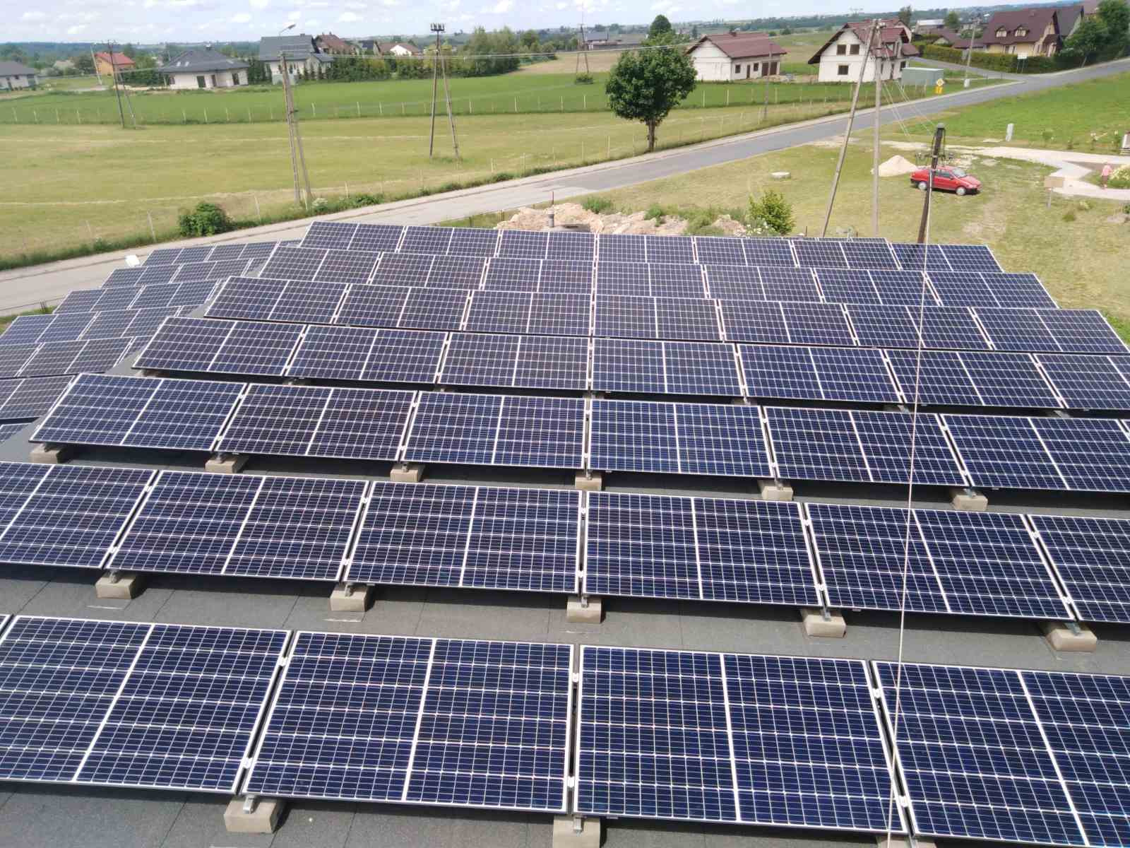 Widok z góry na dużą instalację paneli słonecznych na płaskim dachu, z widocznymi betonowymi podstawami i otaczającym krajobrazem wiejskim z domami i polami.