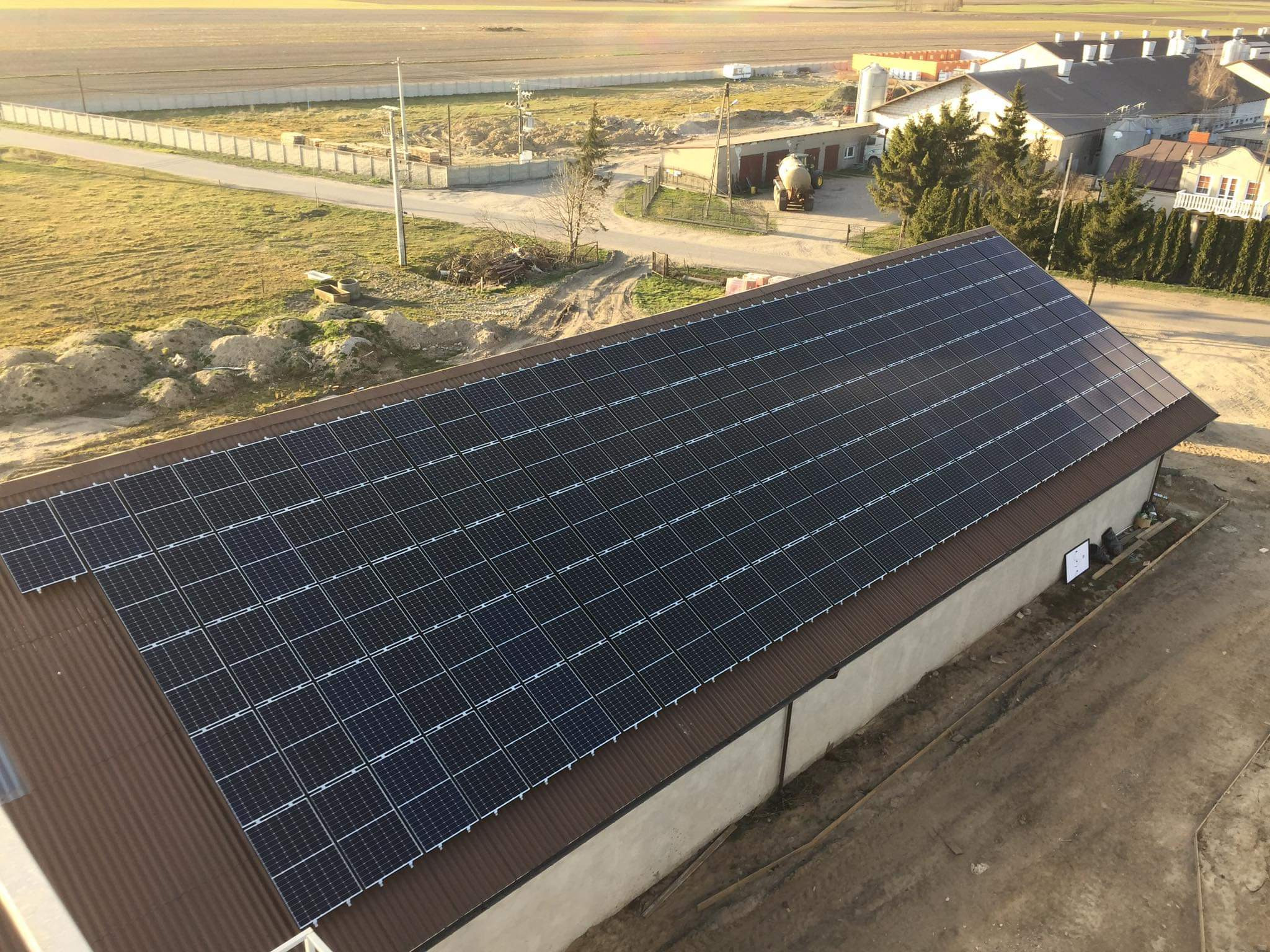 Widok z góry na dużą instalację paneli słonecznych na dachu budynku gospodarczego, na tle wiejskiego krajobrazu o zachodzie słońca.