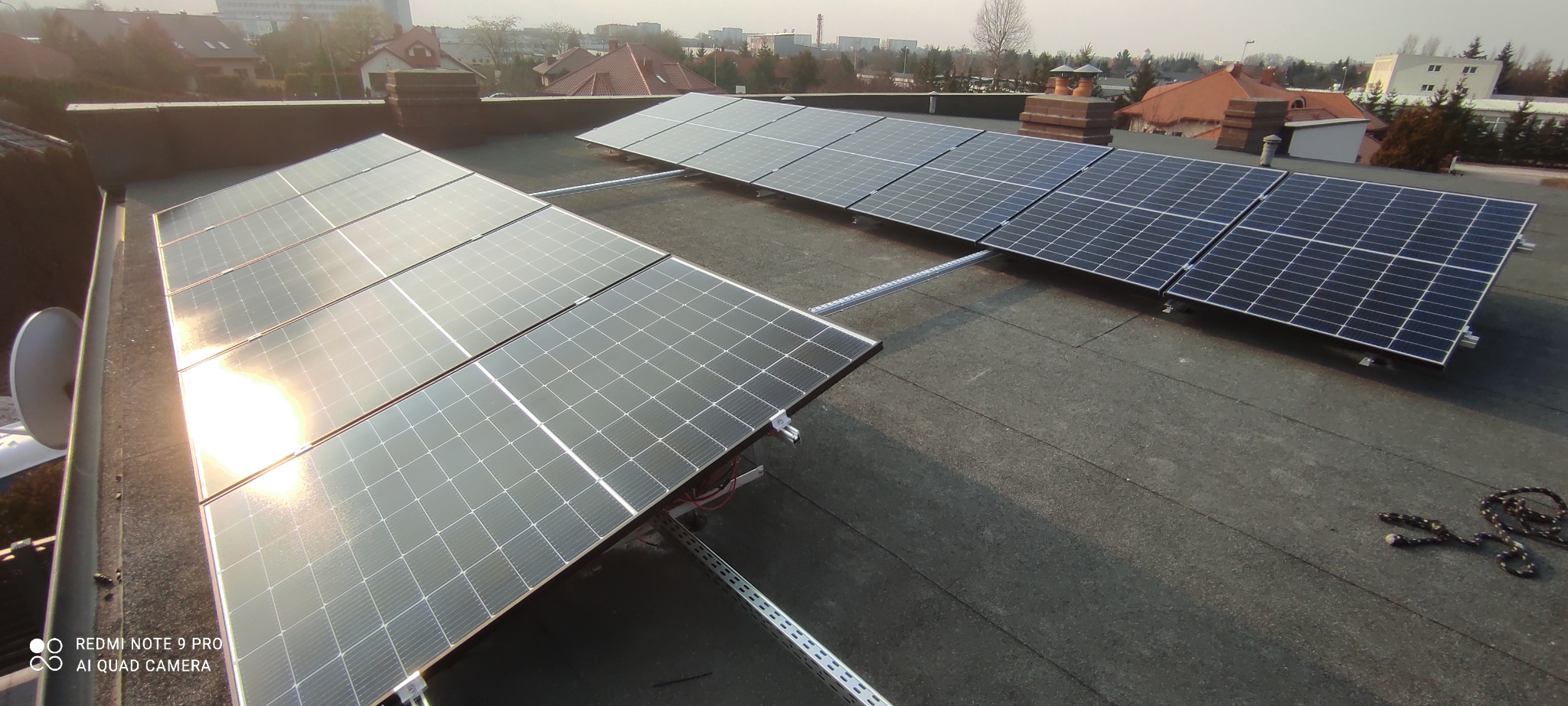 Panele słoneczne zamontowane na płaskim dachu budynku mieszkalnego, widoczne metalowe konstrukcje wsporcze, w tle zabudowa miejska pod lekką mgłą.