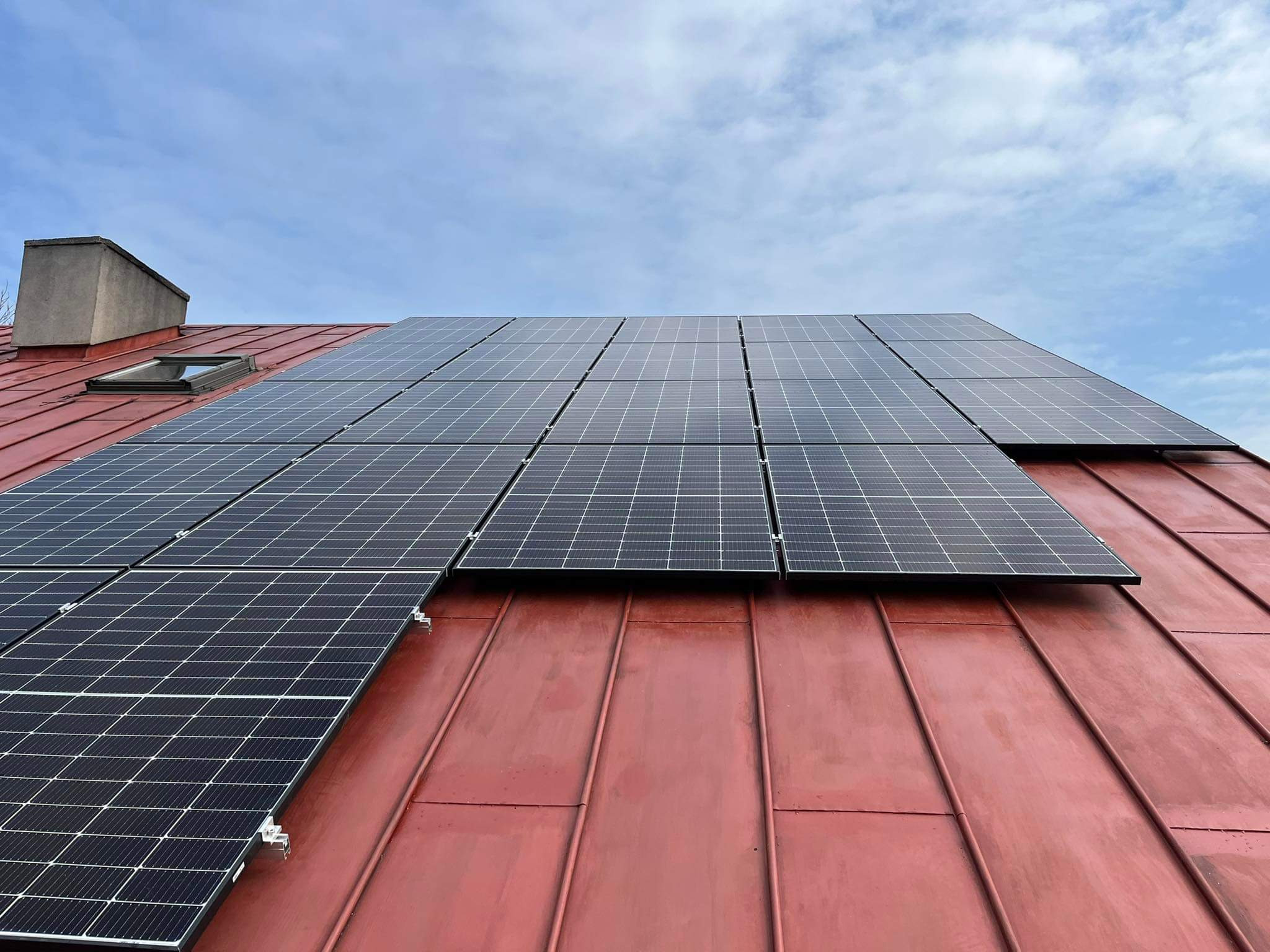Panele słoneczne zamontowane na czerwonym, metalowym dachu z widocznym oknem dachowym i kominem na tle błękitnego nieba z chmurami.