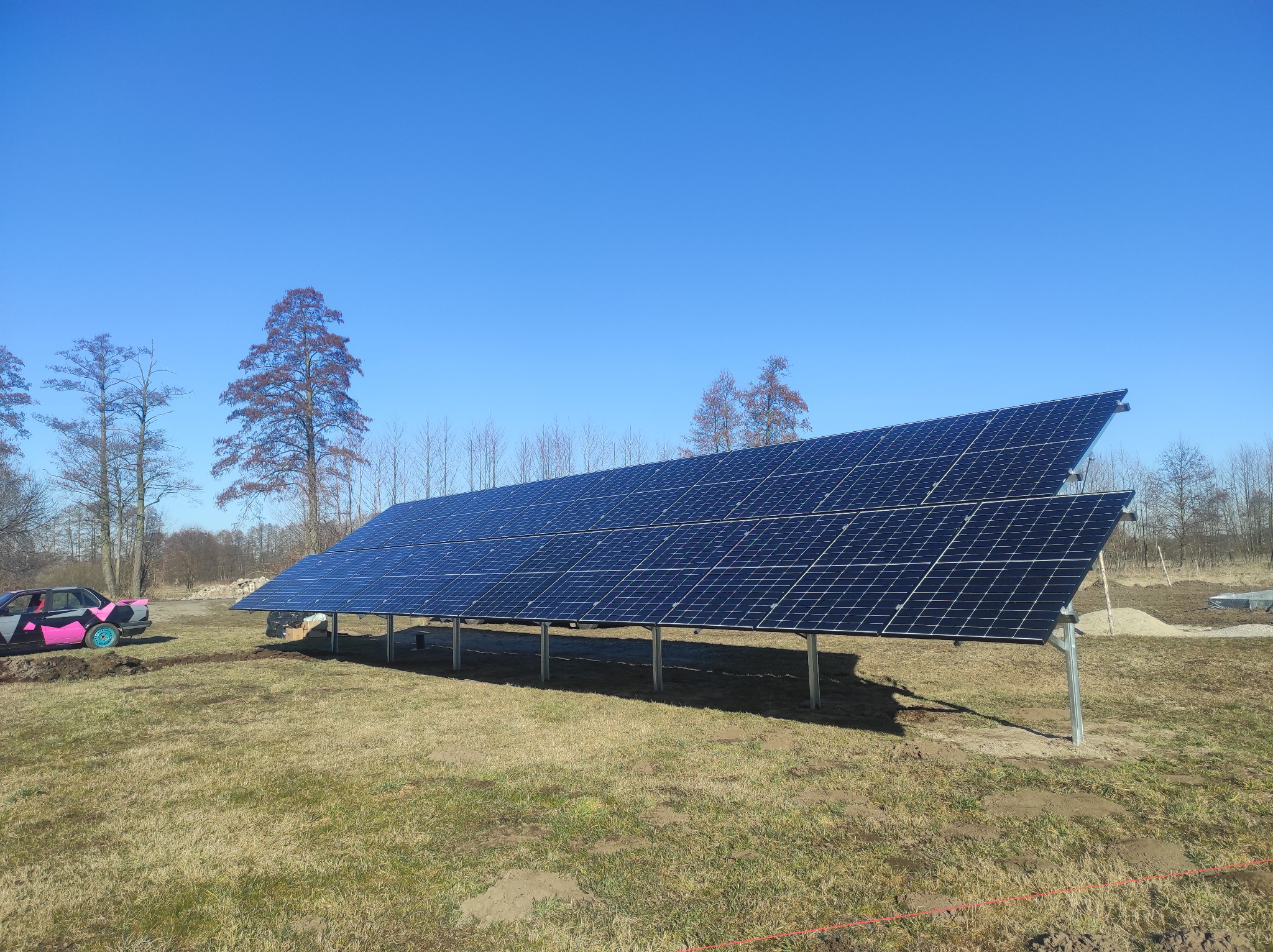 Instalacja paneli słonecznych na metalowej konstrukcji, widoczny na tle błękitnego nieba i drzew, z nietypowym, pomalowanym na różowo i szaro samochodem w tle po lewej stronie.