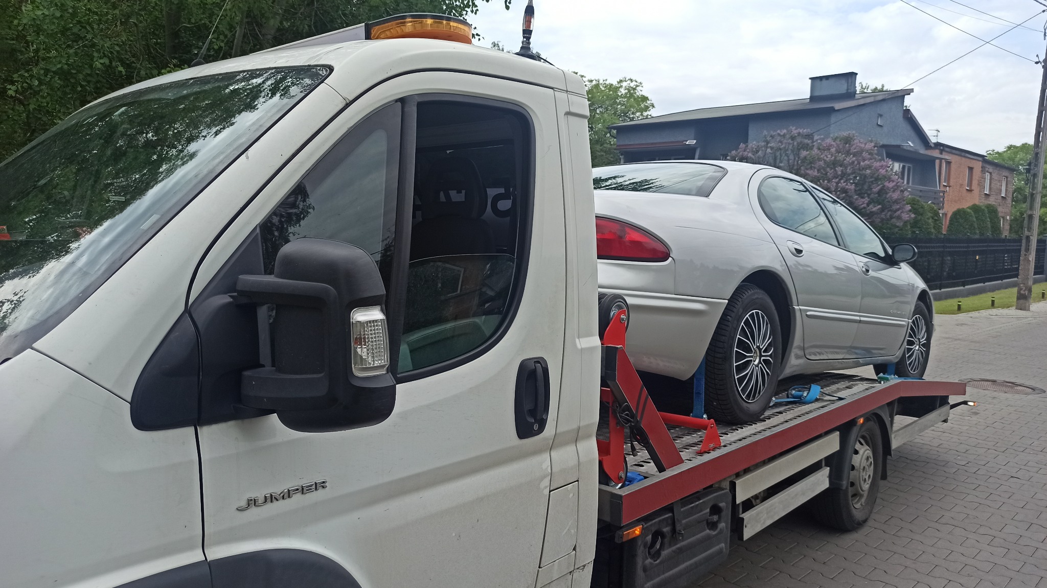 Biała laweta marki Jumper z załadowanym srebrnym sedanem, zabezpieczonym pasami transportowymi, na tle brukowanej ulicy i zabudowań mieszkalnych.