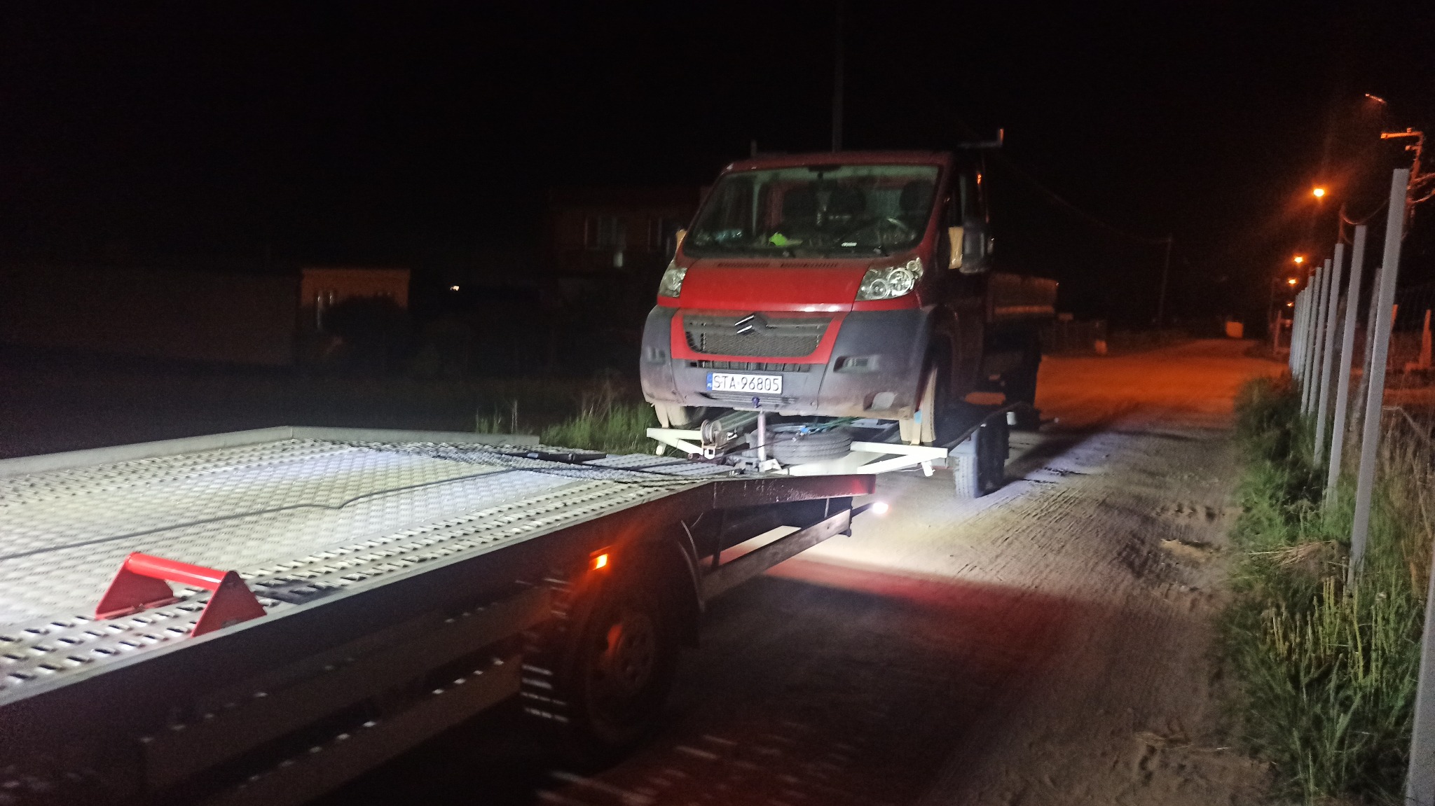 Czerwony samochód dostawczy z uszkodzonym kołem, zabezpieczony na platformie pojazdu holowniczego oświetlonej światłami w nocy.