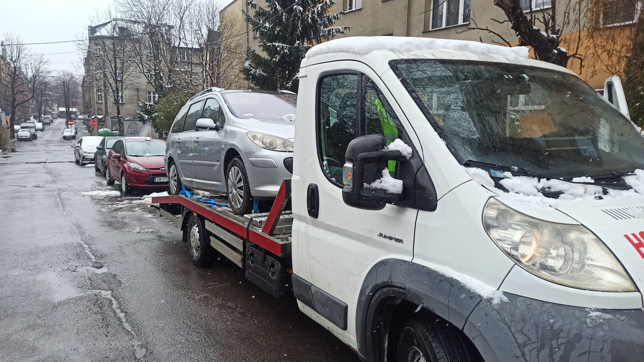 Srebrny samochód osobowy zabezpieczony pasami na platformie białej ciężarówki z kabiną, na tle zimowej ulicy z zaparkowanymi autami i budynkami.