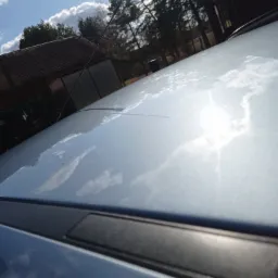 Błyszczący dach jasnego samochodu odbija błękitne niebo z chmurami, widoczny fragment anteny i tło z budynkami oraz drzewami.