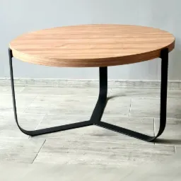 Okrągły stolik kawowy z jasnego drewna na czarnych, metalowych nogach w kształcie litery Y, ustawiony na jasnej płytkowej podłodze przed szarą ścianą.