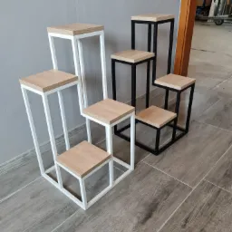 Zestaw sześciu metalowych kwietników o różnej wysokości, z drewnianymi blatami, ustawionych na podłodze z płytek imitujących drewno, na tle szarej ściany i otwartych drzwi.