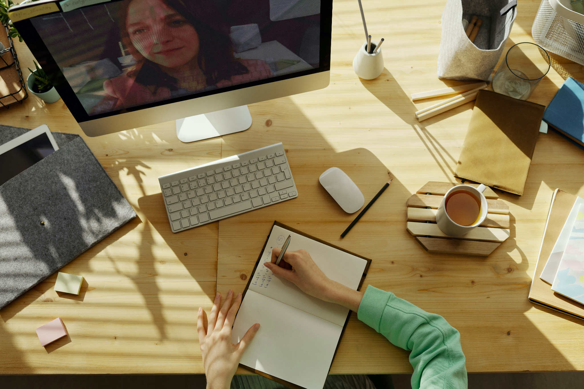 Widok z góry na biurko z komputerem iMac wyświetlającym wideokonferencję, klawiaturą, myszą, notatnikiem, długopisem, kubkiem z herbatą i innymi akcesoriami biurowymi, oświetlone naturalnym światłem.