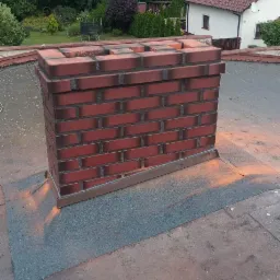 Nowy, ceglany komin z obróbką blacharską na płaskim dachu budynku. Widoczne drzewa w tle, komin w odcieniach czerwieni i brązu.