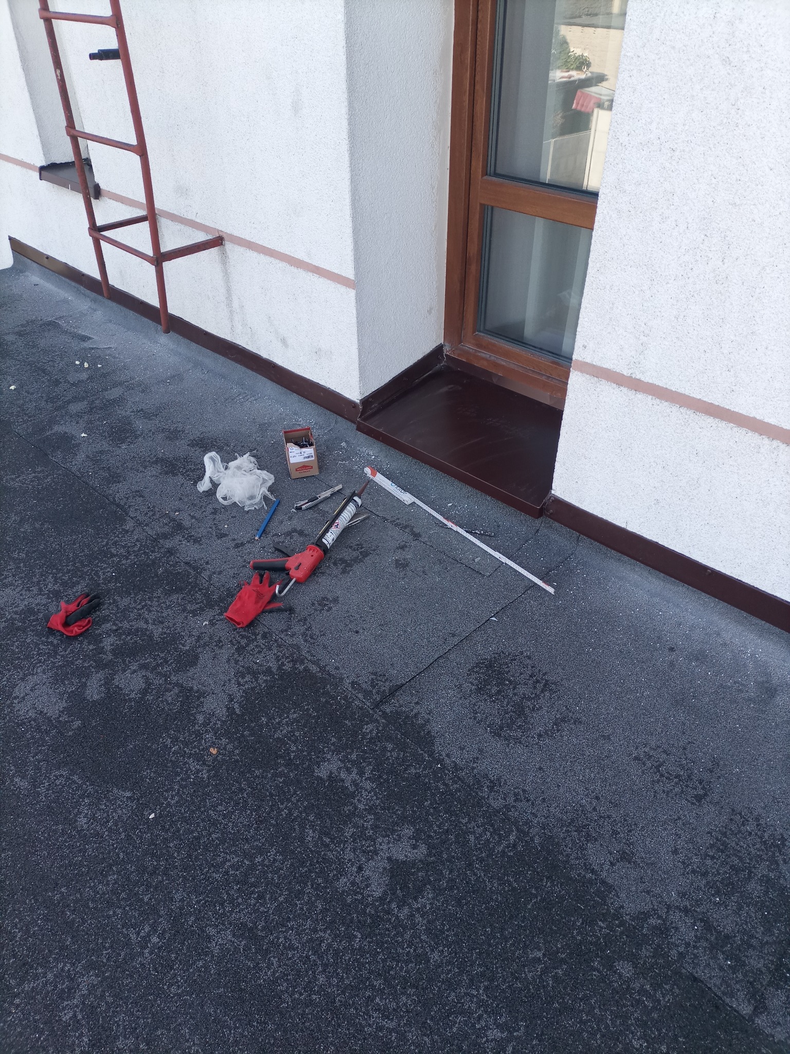 Bitumiczna powierzchnia dachu płaskiego z narzędziami dekarskimi: pistolet do uszczelniacza, miarka, nóż, rękawice i pudełko po wkrętach, widok z góry, drabina oparta o ścianę.