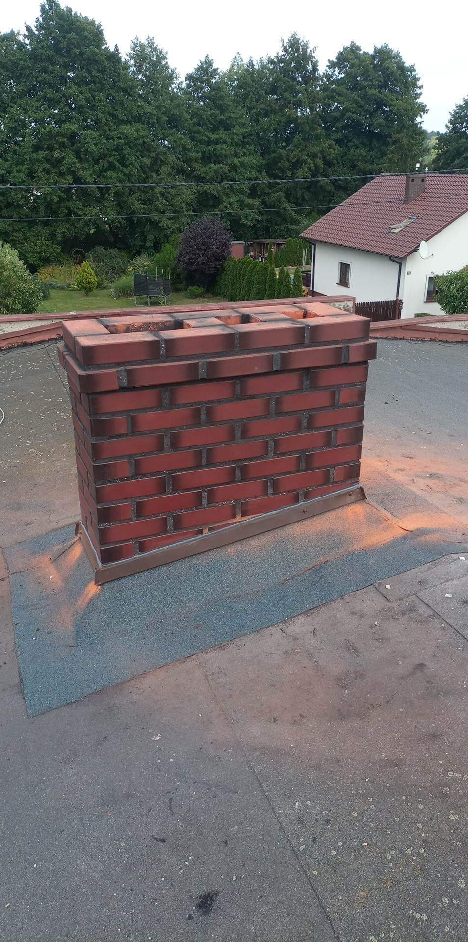 Nowy, ceglany komin z obróbką blacharską na płaskim dachu budynku. Widoczne drzewa w tle, komin w odcieniach czerwieni i brązu.