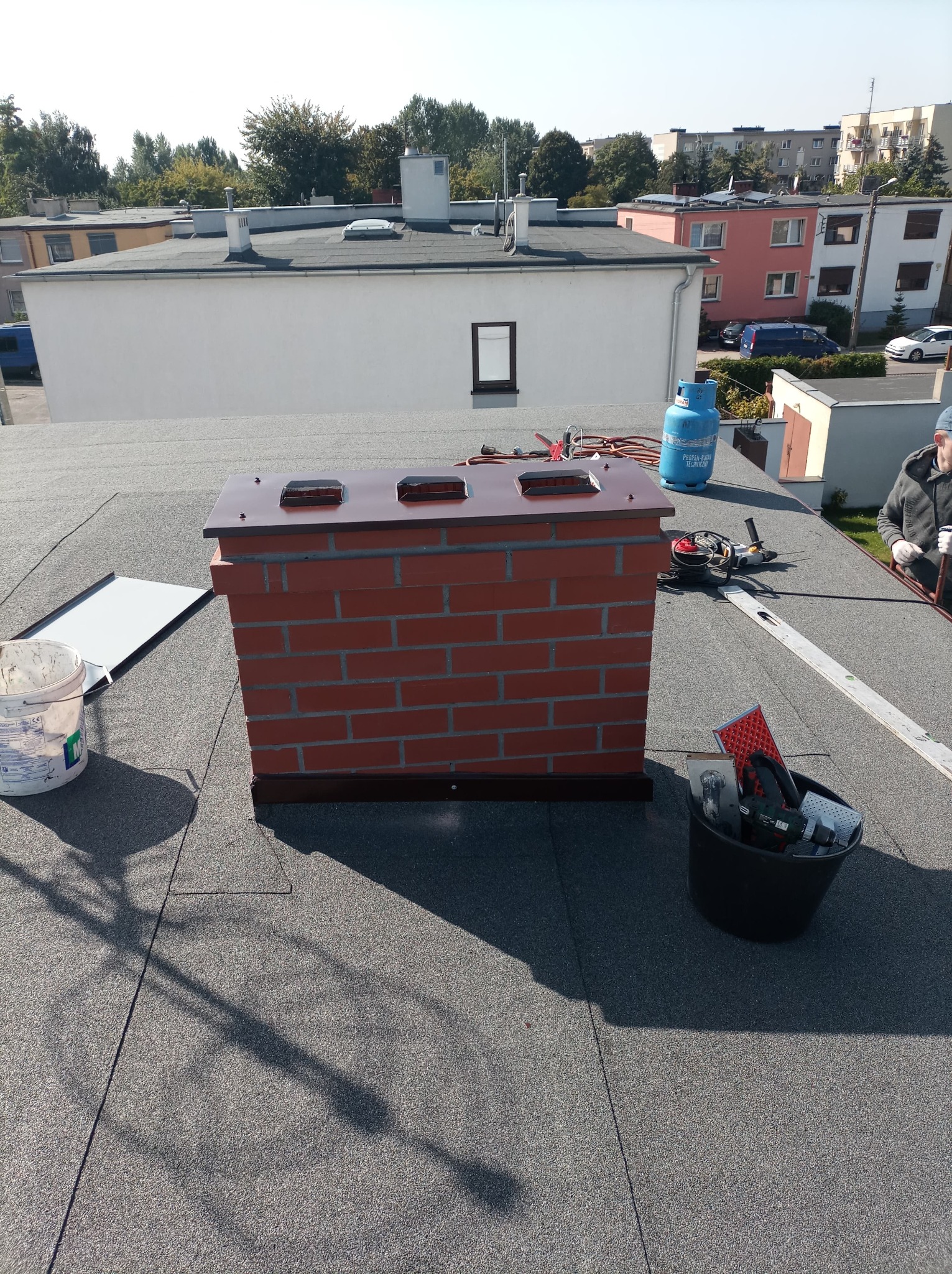 Ceglany komin z metalową obróbką na płaskim dachu pokrytym papą. Widoczne narzędzia, butla gazowa i fragment sylwetki dekarza w tle.