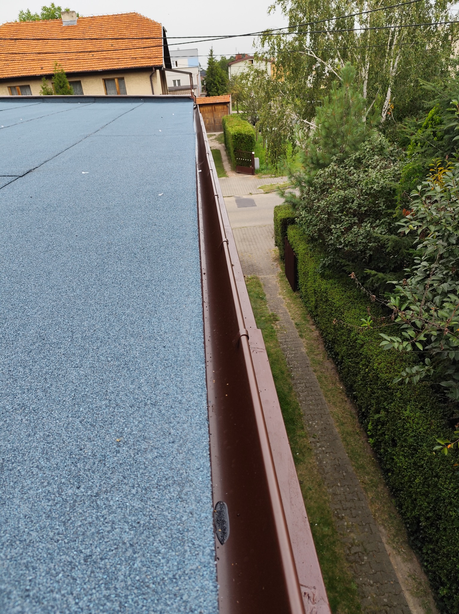 Niebieski dach pokryty papą z brązową rynną wzdłuż krawędzi. Widok z góry na dach i przydomowy ogródek z zielonymi krzewami i drzewami.