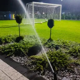 Nocne nawadnianie trawnika z automatycznym systemem zraszaczy, w tle bramka do piłki nożnej i krzewy ozdobne.