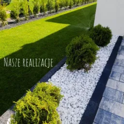 Ogród z równo przystrzyżonym trawnikiem, ozdobnymi krzewami iglastymi i białymi kamieniami, w tle plac zabaw dla dzieci.