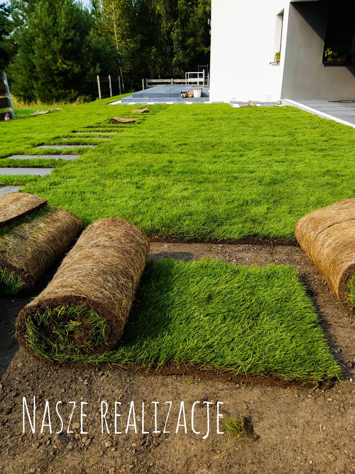 Układanie trawy z rolki na nowo zagospodarowanym terenie przy nowoczesnym domu. Widoczne rolki trawy i fragmenty już ułożonej darni, z elementami ścieżki z płyt betonowych.