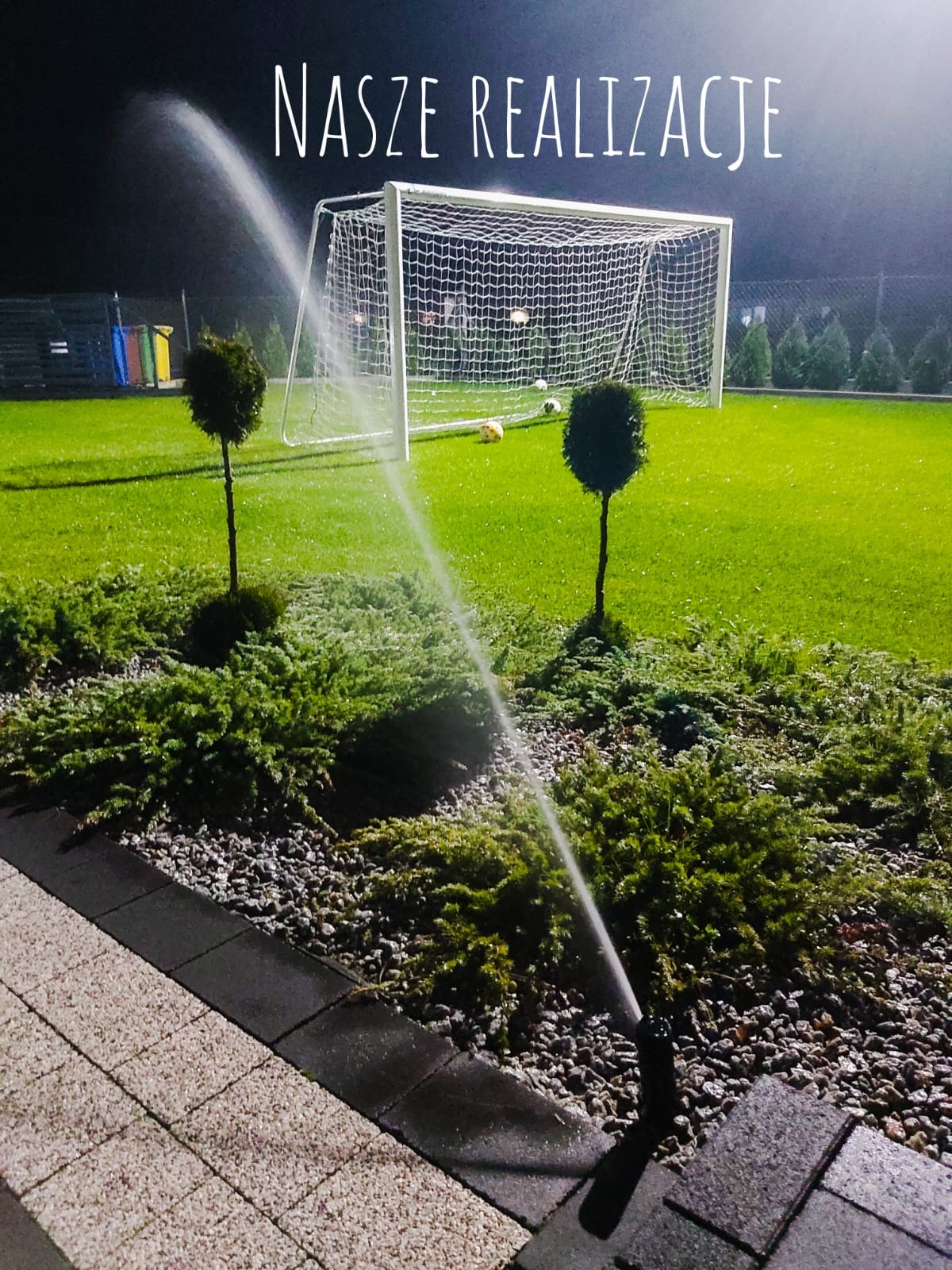 Nocne nawadnianie trawnika z automatycznym systemem zraszaczy, w tle bramka do piłki nożnej i krzewy ozdobne.