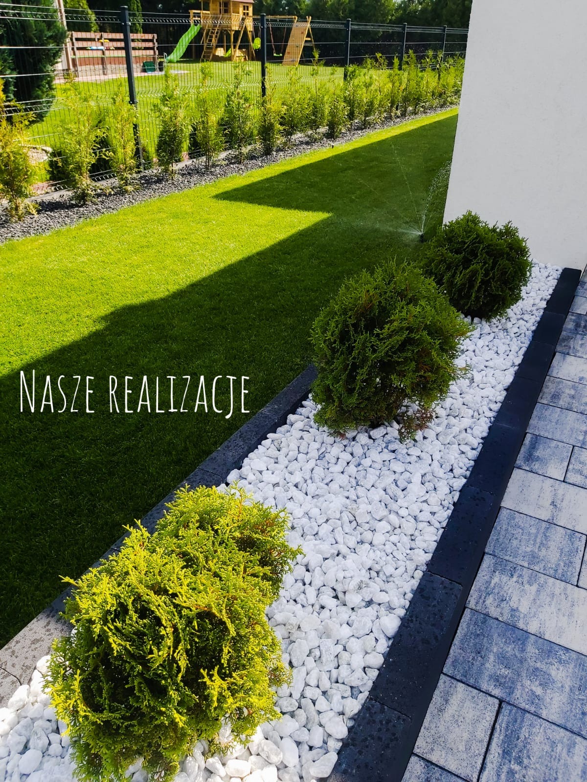 Ogród z równo przystrzyżonym trawnikiem, ozdobnymi krzewami iglastymi i białymi kamieniami, w tle plac zabaw dla dzieci.