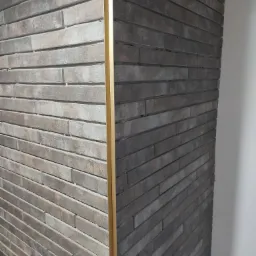 Fragment elewacji wykończonej klinkierem w dwóch odcieniach szarości, połączonych złotą listwą dekoracyjną, widoczny narożnik budynku.